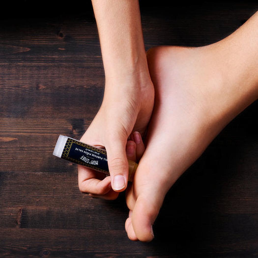 A hand holds Moms Stuff Salve All Purpose Pinon Salve - Large Tube on a dark wooden surface next to a foot.
