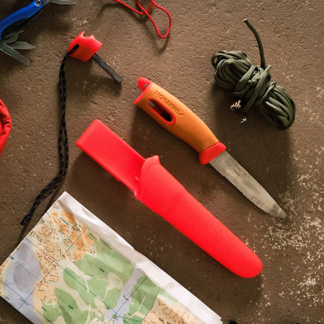 A top-down view of the Morakniv Companion Spark Emergency knife in Red with its red sheath, a map, and green rope on a brown surface.