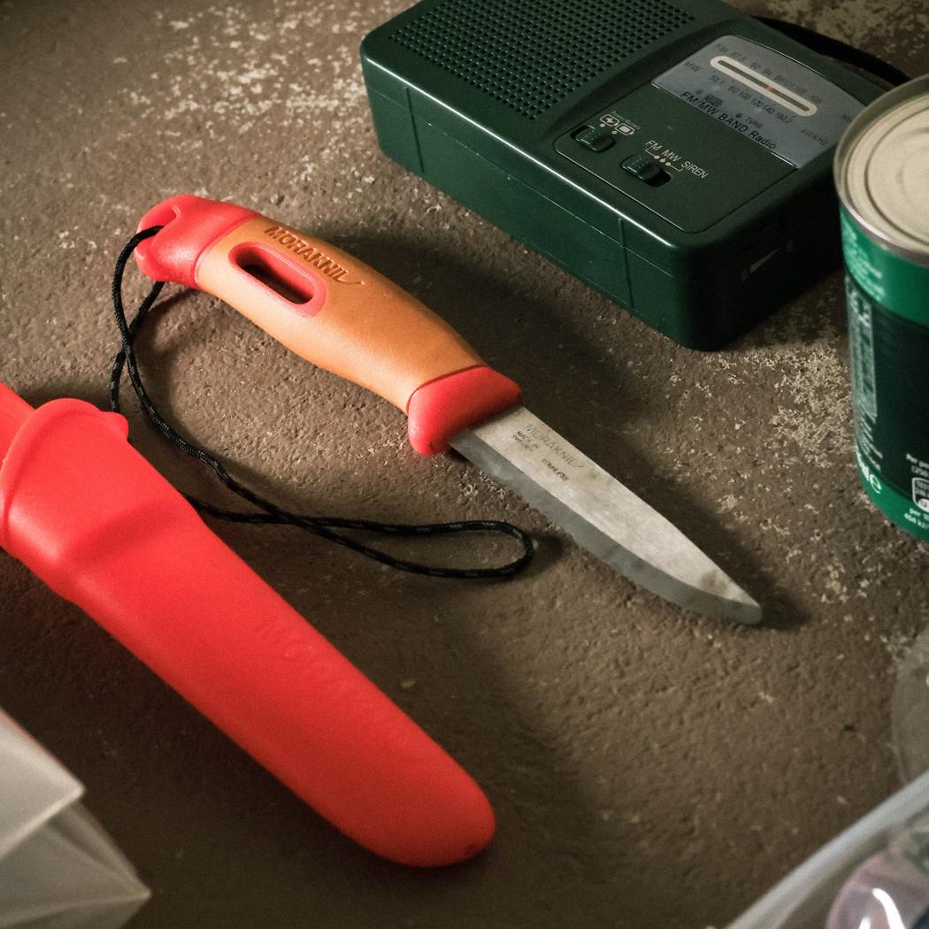 A red Morakniv Companion Spark Emergency knife with its sheath lies on a concrete surface next to a green radio and a can.