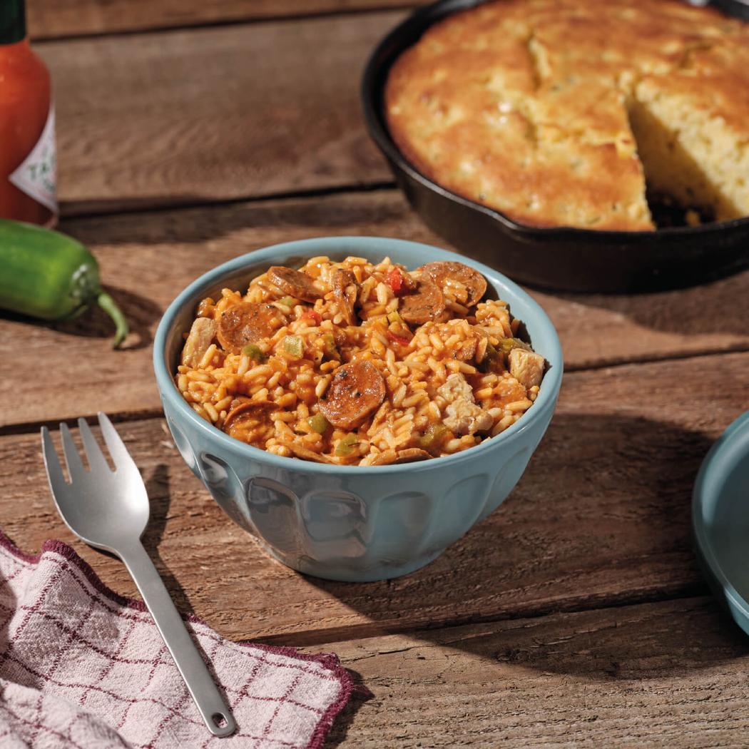 A bowl of Mountain House Cajun Style Jambalaya sits on a wooden table with a fork and a skillet of cornbread in the background.
