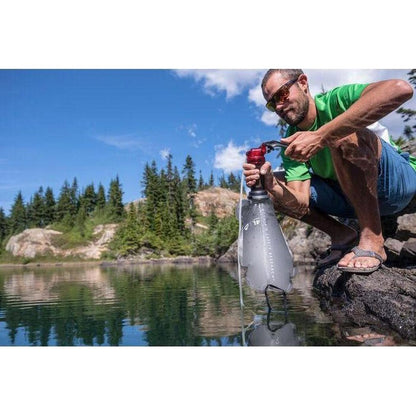 A man crouches by a lake using an MSR DromLite 2L bag to fill it with water, surrounded by trees and rocky terrain.