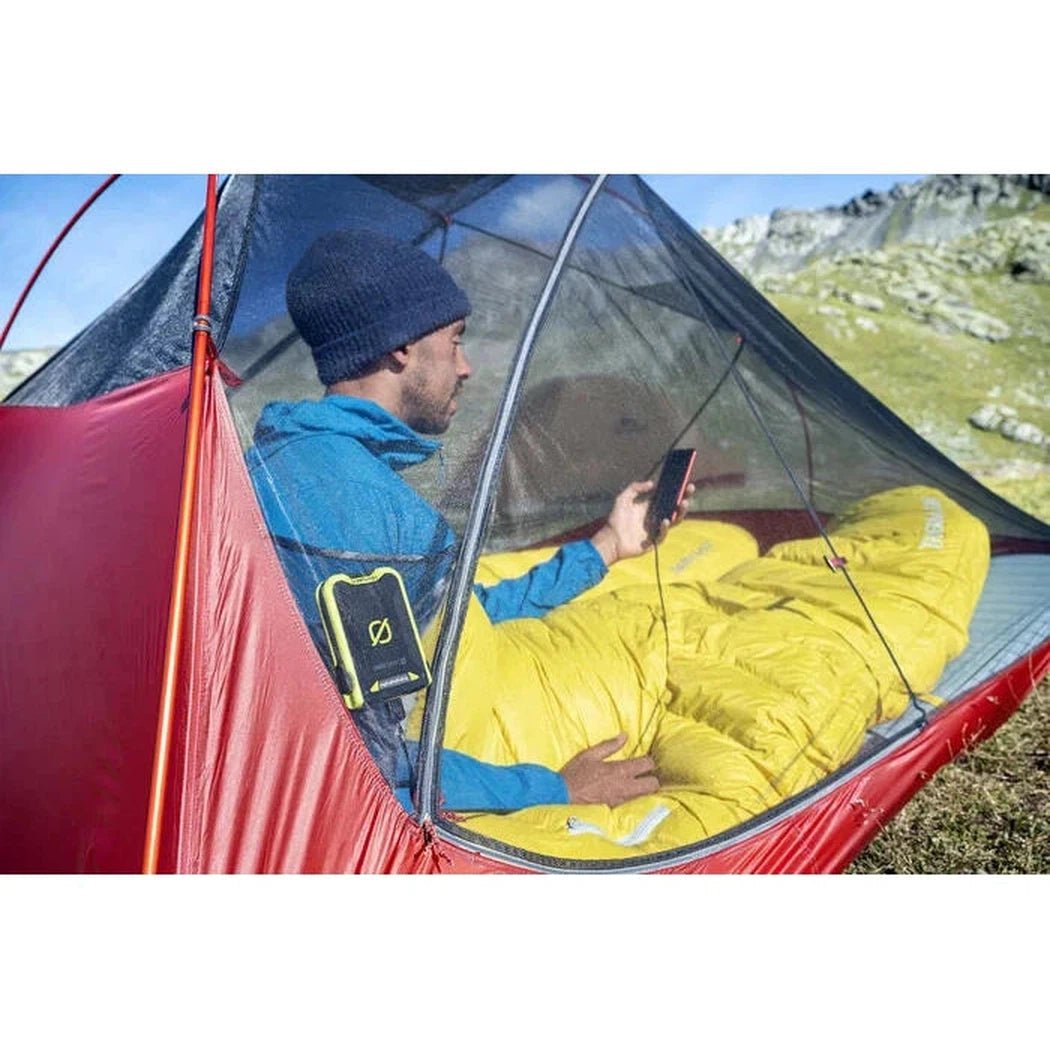 A man in a blue jacket sits in a red MSR FreeLite 2 Tent with a yellow sleeping bag, using a phone, against a mountainous backdrop.