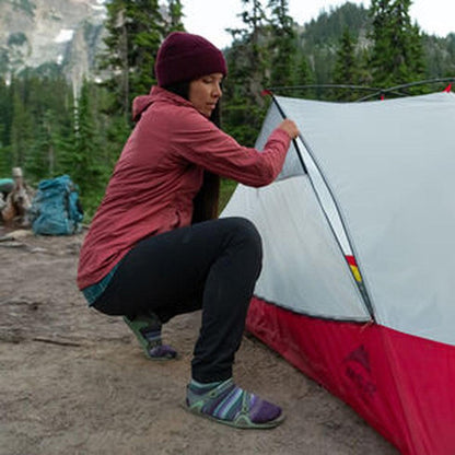 A woman crouches to adjust the MSR Hubba Hubba 2 Tent V9, which has a red and gray exterior, in a forested mountain setting.