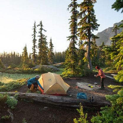 A person sets up the MSR Hubba Hubba 2 Tent V9 in a forested campsite with tall trees and a mountain in the background.