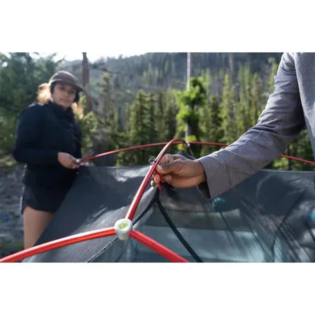 A person holds a red pole of the MSR Hubba Hubba LT 2P Tent while another stands by a forested mountain backdrop.