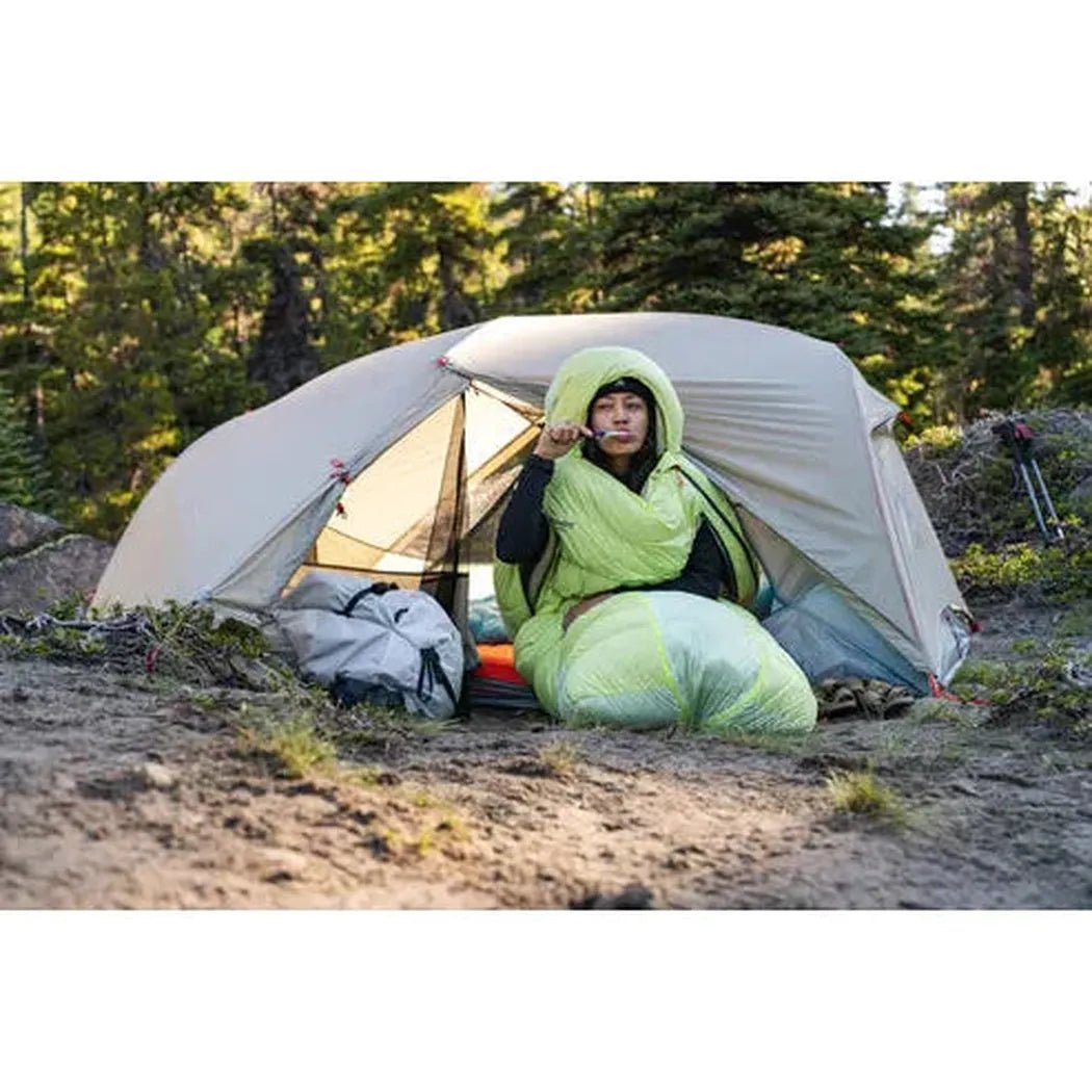 A person wearing a green sleeping bag sits inside the MSR Hubba Hubba LT 2P Tent in a forested area with a gray backpack nearby.