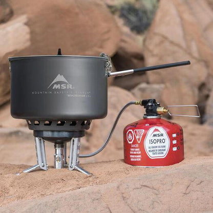 A charcoal MSR WindBurner Group Stove System sits on rocky ground with its red canister.