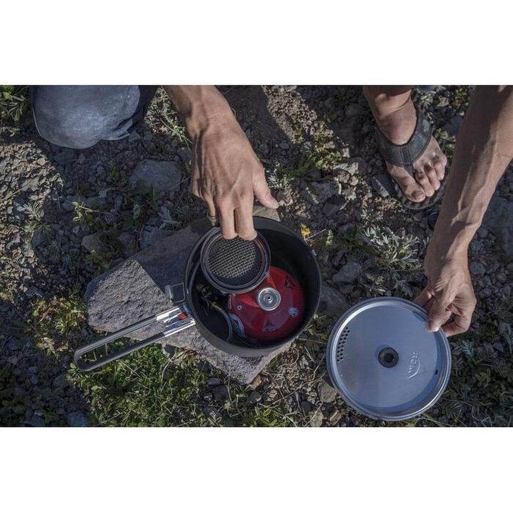 A person kneels on rocky ground holding the lid of the MSR WindBurner Group Stove System while the red stove sits in a pot on a rock.