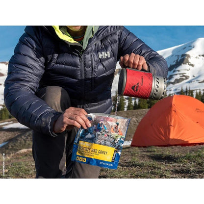 A person in a charcoal jacket pours from a red MSR WindBurner stove into a package of Viscous and Gravy while camping near an orange tent.