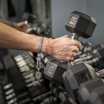 A hand wearing a blue Nite Ize Cinch-A-Lot Stretch Strap holds a 20-pound dumbbell in a gym setting.