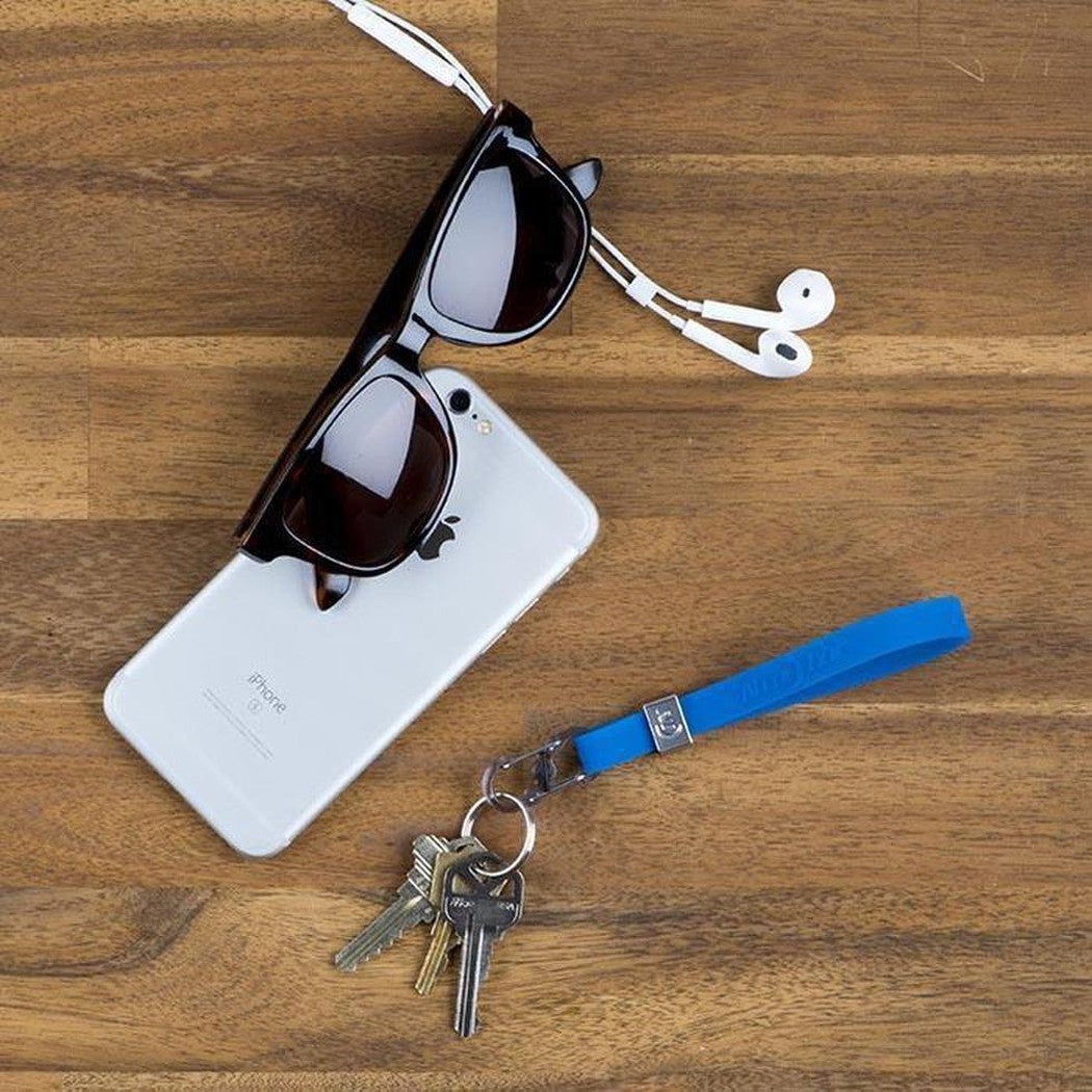 A top-down view of a blue Nite Ize Cinch-A-Lot Stretch Strap with keys attached, resting on a wooden surface next to a smartphone and sunglasses.