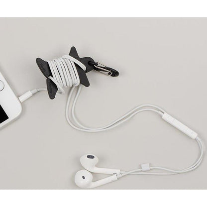 A black Nite Ize Curvyman Cord Supervisor holds a white cord, with white earbuds and a phone visible on a light background.
