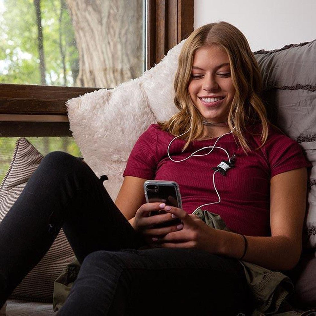 A woman wearing the black Nite Ize Curvyman Cord Supervisor sits on a couch with a phone, smiling, while looking out a window.