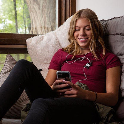 A woman wearing the black Nite Ize Curvyman Cord Supervisor sits on a couch with a phone, smiling, while looking out a window.