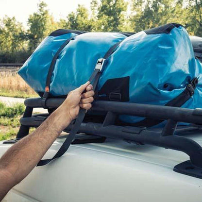 A hand secures a 12ft Nite Ize Dual CamJam Tie Down System strap around blue gear on a vehicle roof rack, with trees in the background.