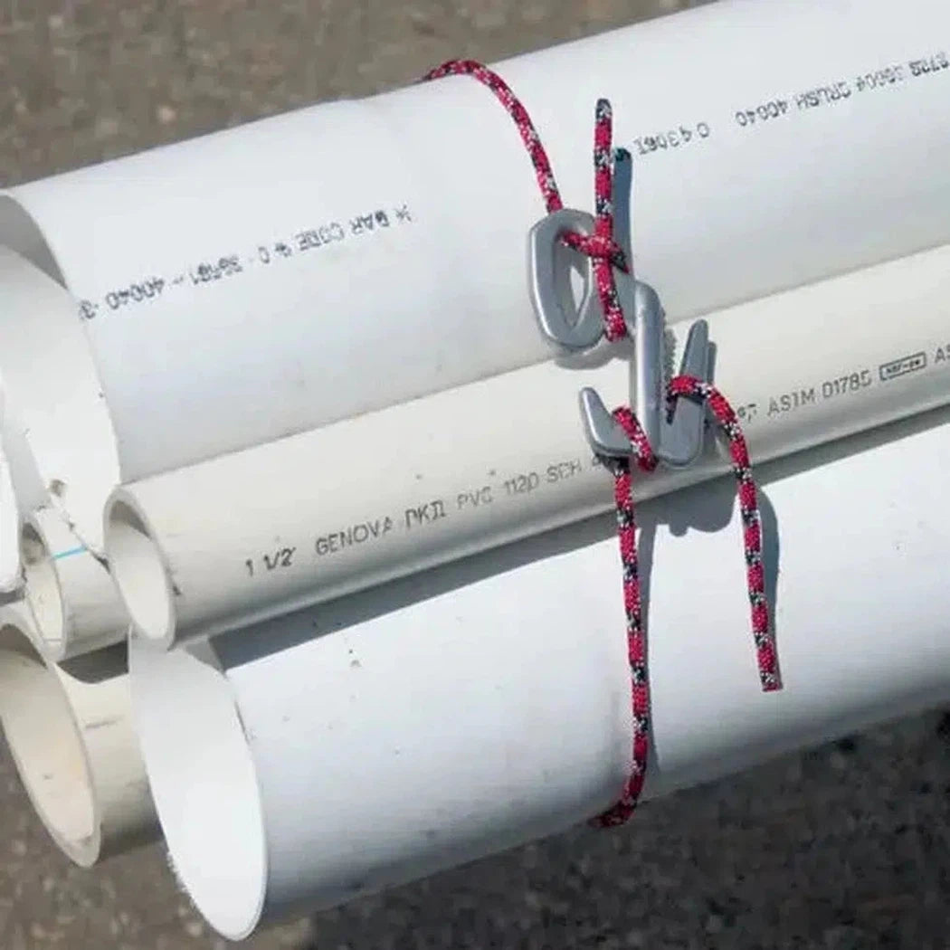 A close-up of the Nite Ize Figure 9 Rope Tightener - Small in Silver, showing it securing white PVC pipes with a red and black rope against a gravel background.