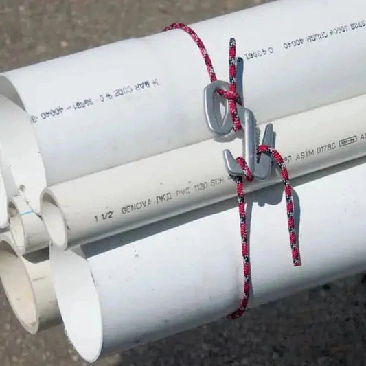 A close-up of the Nite Ize Figure 9 Rope Tightener - Small in Silver, showing it securing white PVC pipes with a red and black rope against a gravel background.