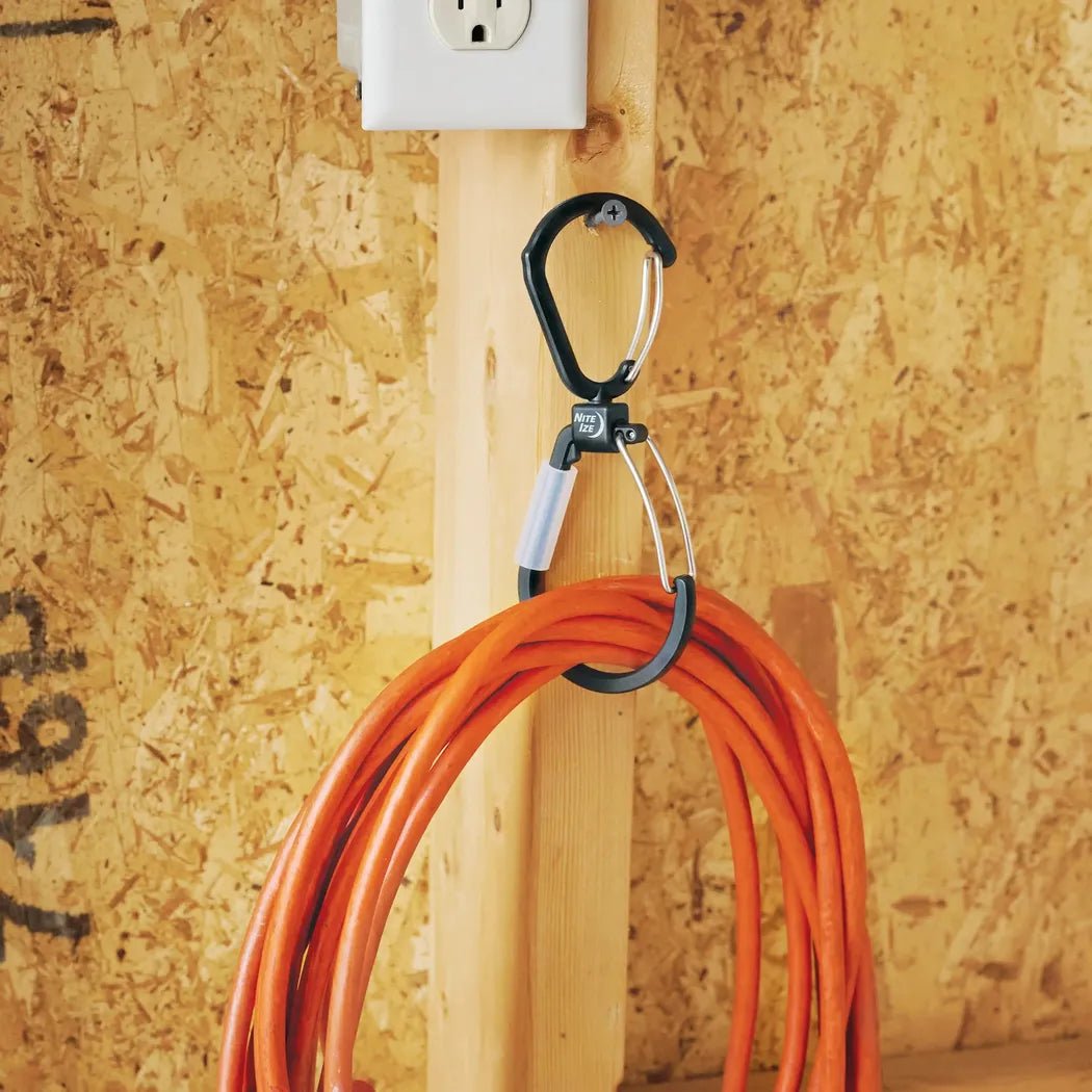 A black Nite Ize FlipOut Dual Carabiner hangs an orange extension cord from a wooden beam with a power outlet above.