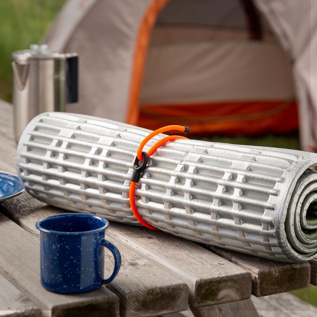A rolled camping mat on a wooden table with a blue mug and tent in the background, secured with a bright orange Nite Ize Gear Tie Loopable Twist Tie 24 inch - 2 Pack.