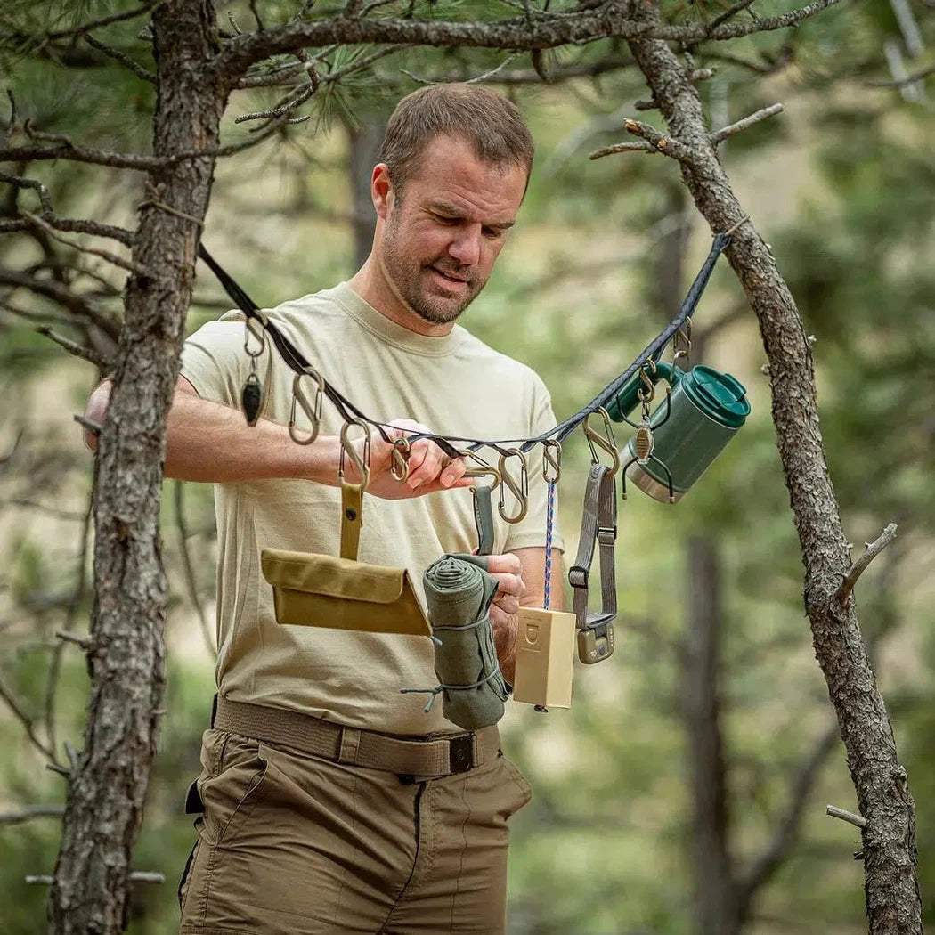 A man in a forest uses the Nite Ize GearLine Organization System with tactical gear hanging from tree branches.