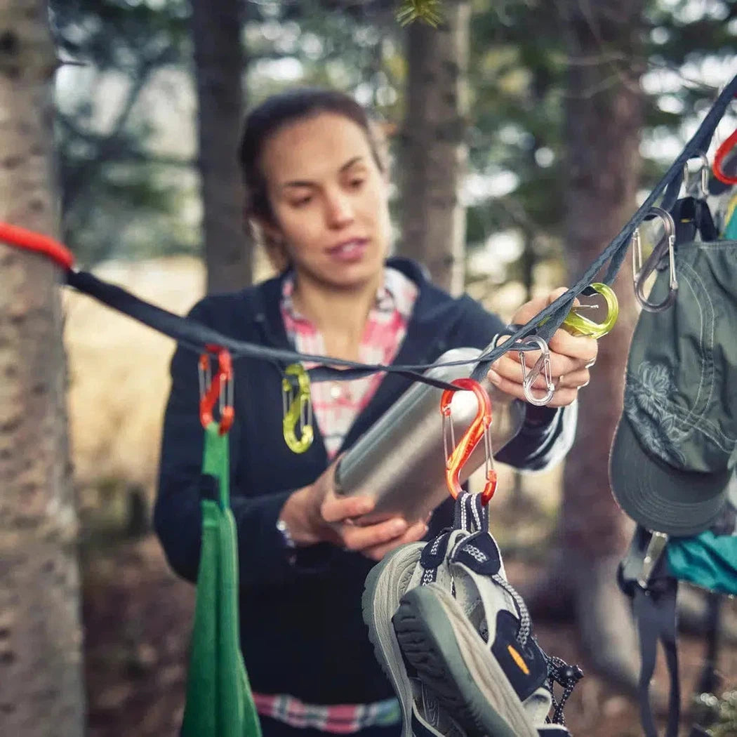A woman in a forest uses the Nite Ize GearLine Organization System to hang a water bottle and shoes.