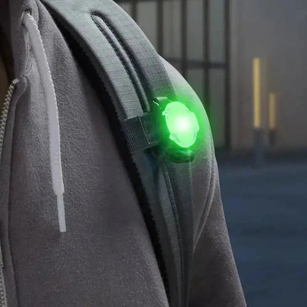 A close-up of the Nite Ize GoLit Rechargeable Visibility Light - Disc-O Tech attached to a gray jacket strap, glowing green in a dimly lit outdoor setting.