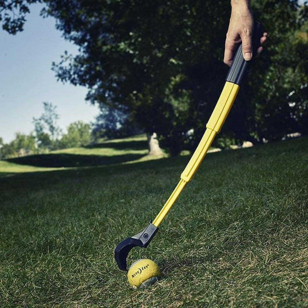 A hand holds the yellow Nite Ize Huck 'n Tuck Collapsible Thrower over a yellow ball on grass with trees in the background.