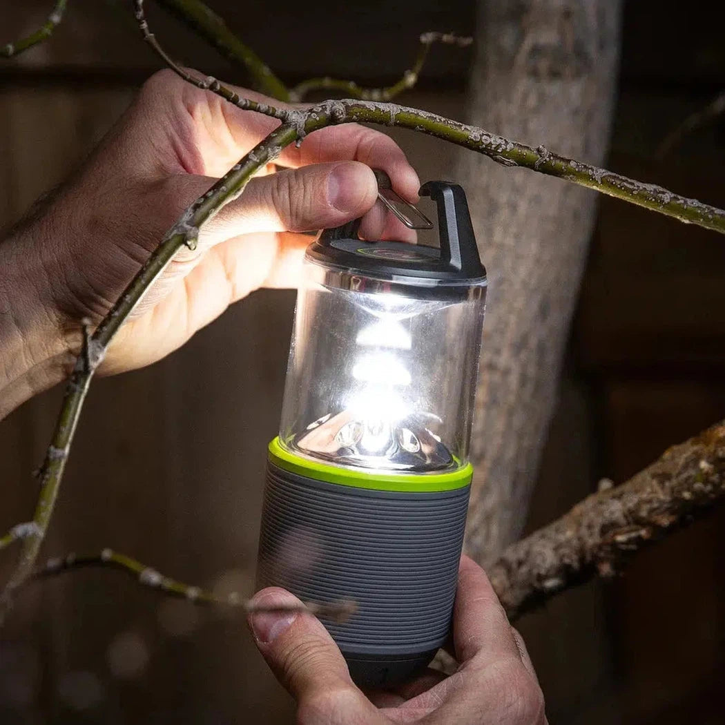 A hand holds the Nite Ize Radiant RL2 Rechargeable Lantern with a bright light on, against tree branches.