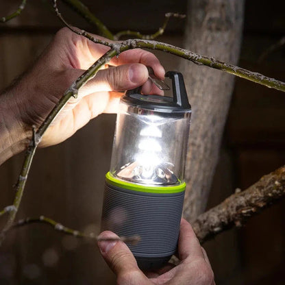 A hand holds the Nite Ize Radiant RL2 Rechargeable Lantern with a bright light on, against tree branches.