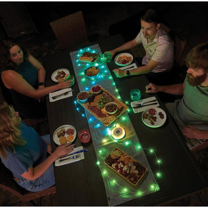 A top-down view of a table with Nite Ize Radiant StarLit Rechargeable Lantern and String Light DiscO in green lighting, showing plates of food and people eating.