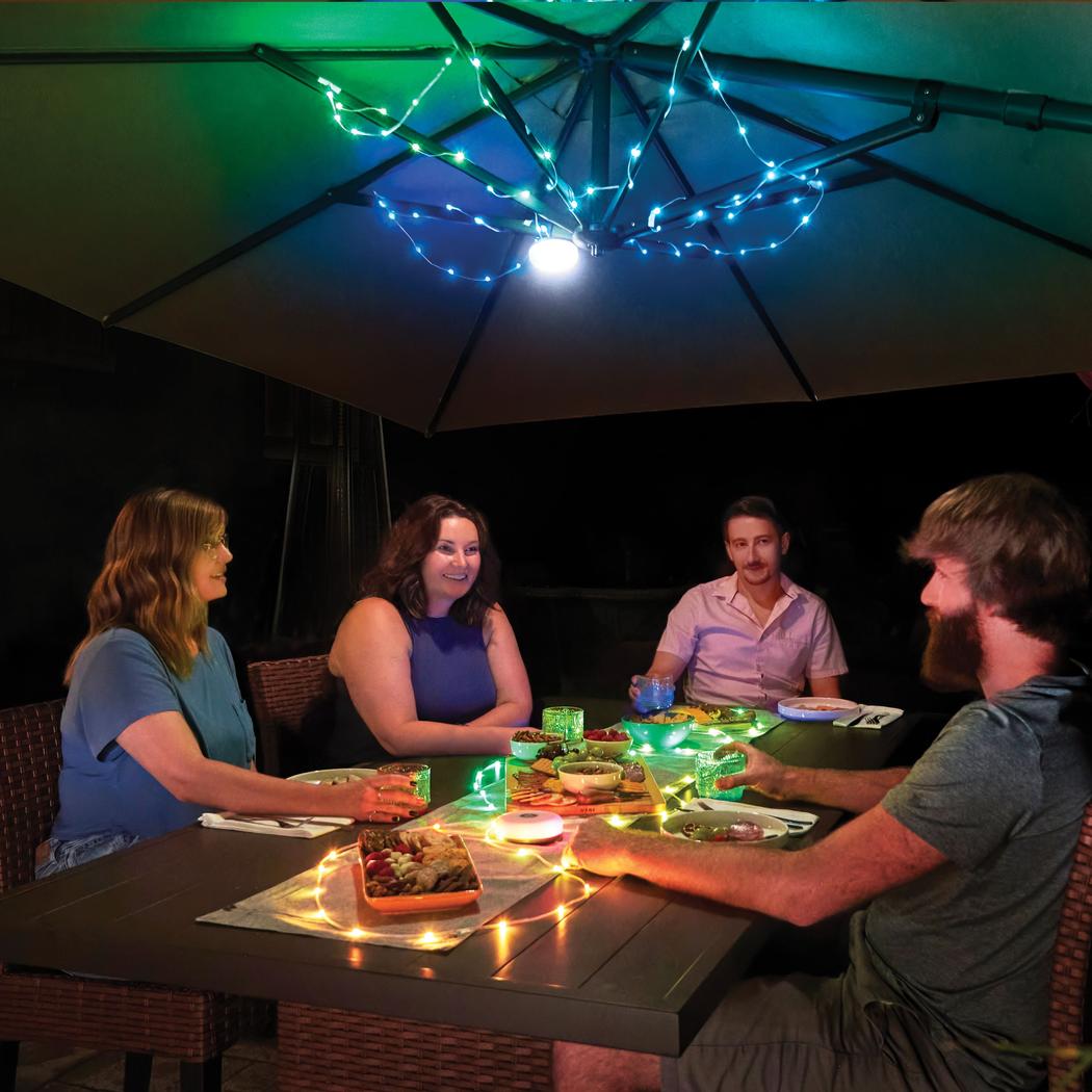 Four people dine under a large umbrella decorated with Nite Ize Radiant StarLit Rechargeable Lantern and String Light DiscO lights at night.