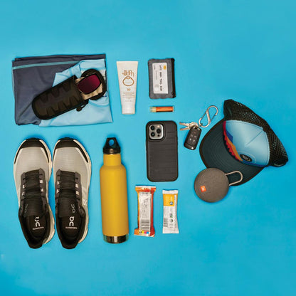 A top-down view of the Nite Ize RunOff Waterproof 12L Backpack in Gray, showing its contents including shoes, a phone, and a hat on a blue background.