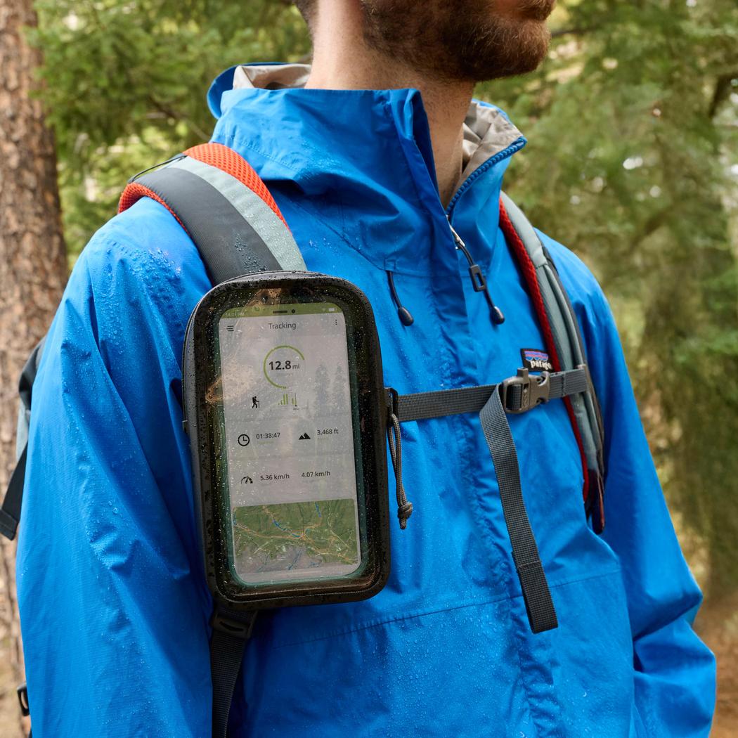 A man in a blue jacket wears the black Nite Ize RunOff Waterproof Phone Case Plus on his backpack strap, with a forest background.