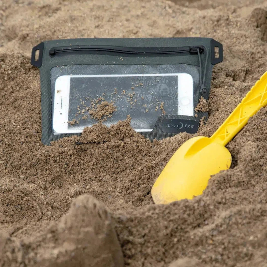 A yellow shovel and a white phone in a charcoal Nite Ize RunOff Waterproof Pocket lie in the sand.