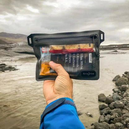 A hand holds the Nite Ize RunOff Waterproof Pocket over a rocky riverbank with mountains in the distance.