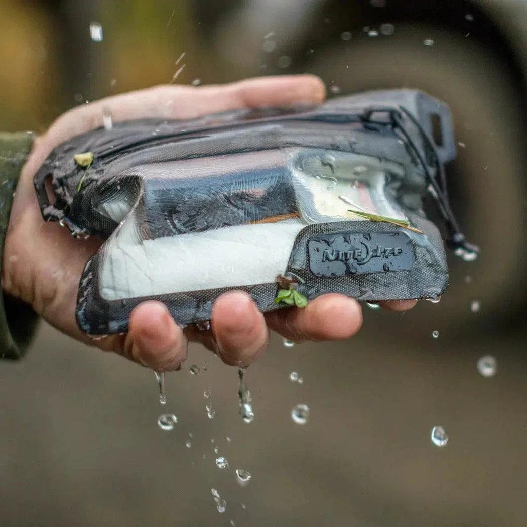 A hand holds the Nite Ize RunOff Waterproof Wallet as water drips from it, showing its transparent front and zipper.