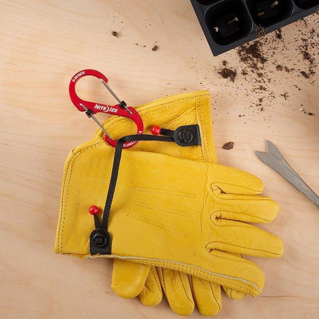 A pair of yellow gloves with a red Nite Ize S-Biner Slidelock Aluminum #4 carabiner attached, on a light wooden surface with a seedling tray and soil nearby.