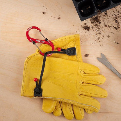 A pair of yellow gloves with a red Nite Ize S-Biner Slidelock Aluminum #4 carabiner attached, on a light wooden surface with a seedling tray and soil nearby.