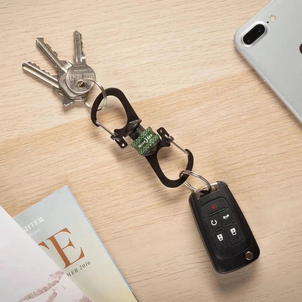 A top-down view of the Nite Ize SlideLock 360 Magnetic Locking Dual Carabiner in Charcoal/Graphite holding keys and a car key fob on a light wood surface with a phone nearby.