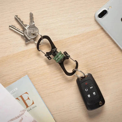 A top-down view of the Nite Ize SlideLock 360 Magnetic Locking Dual Carabiner in Charcoal/Graphite holding keys and a car key fob on a light wood surface with a phone nearby.