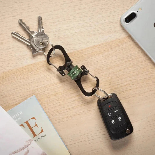 A top-down view of the Nite Ize SlideLock 360 Magnetic Locking Dual Carabiner in Charcoal/Graphite holding keys and a car key fob on a light wood surface with a phone nearby.