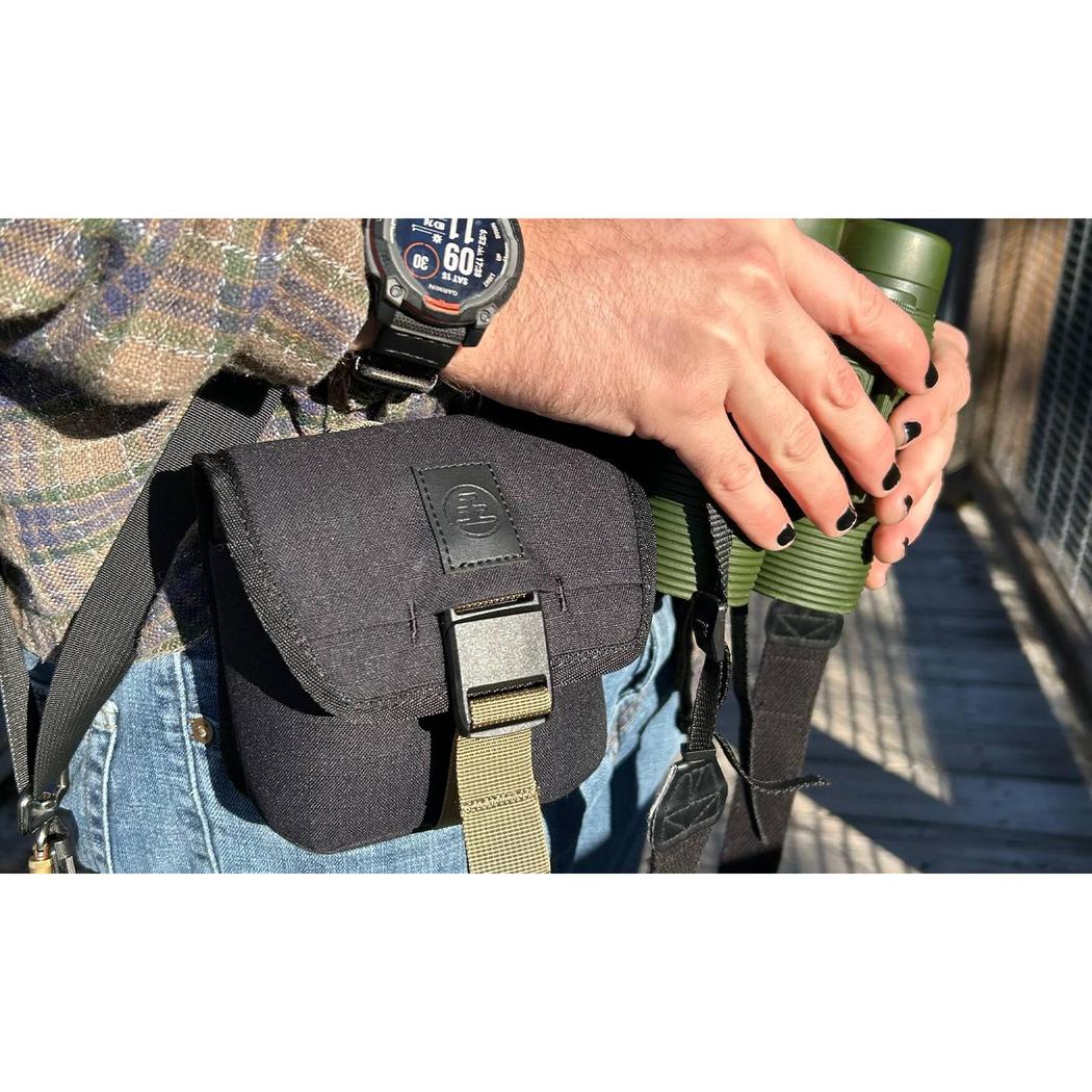 A person wearing blue jeans and a plaid shirt holds green binoculars while a Charcoal Nocs Provisions Standard Issue Pouch hangs at their waist.