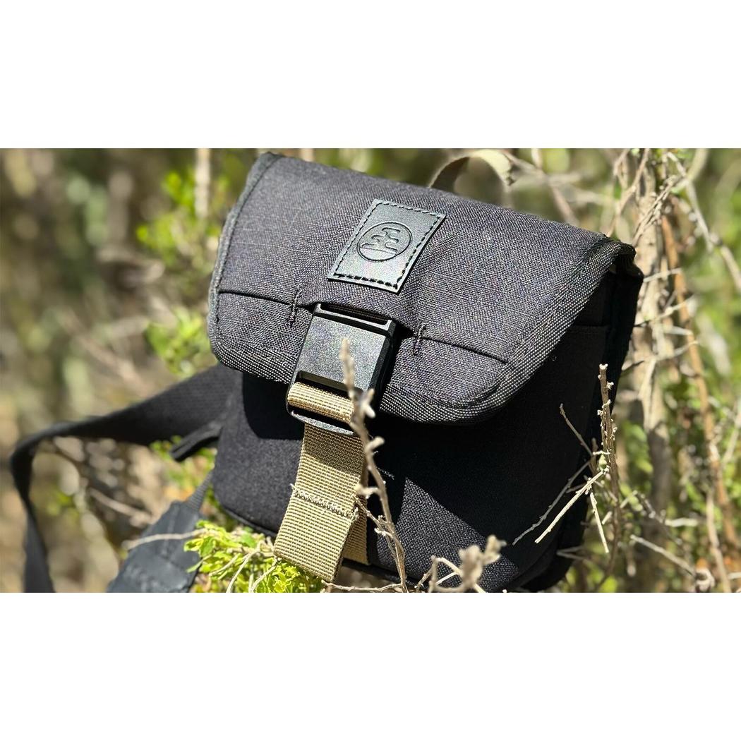 A charcoal Nocs Provisions Standard Issue Pouch with a tan buckle strap sits outdoors among dry grass and green foliage.