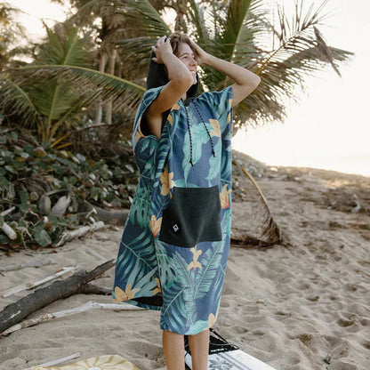 A model wears the Nomadix Changing Poncho in Agua Blue on a sandy beach with palm trees in the background.