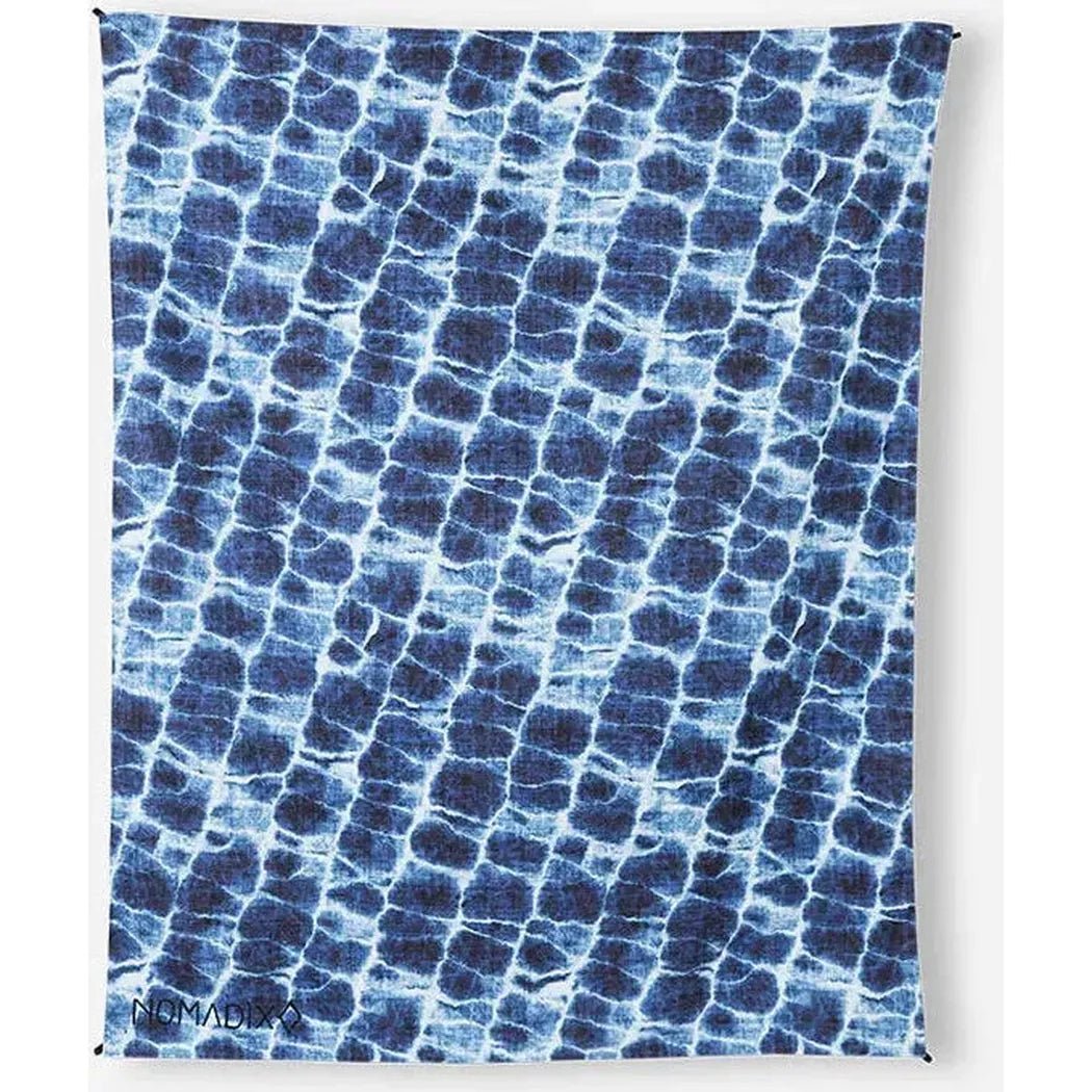 A Nomadix Festival Blanket in Baja Aqua with a blue and white pattern hangs against a white background.