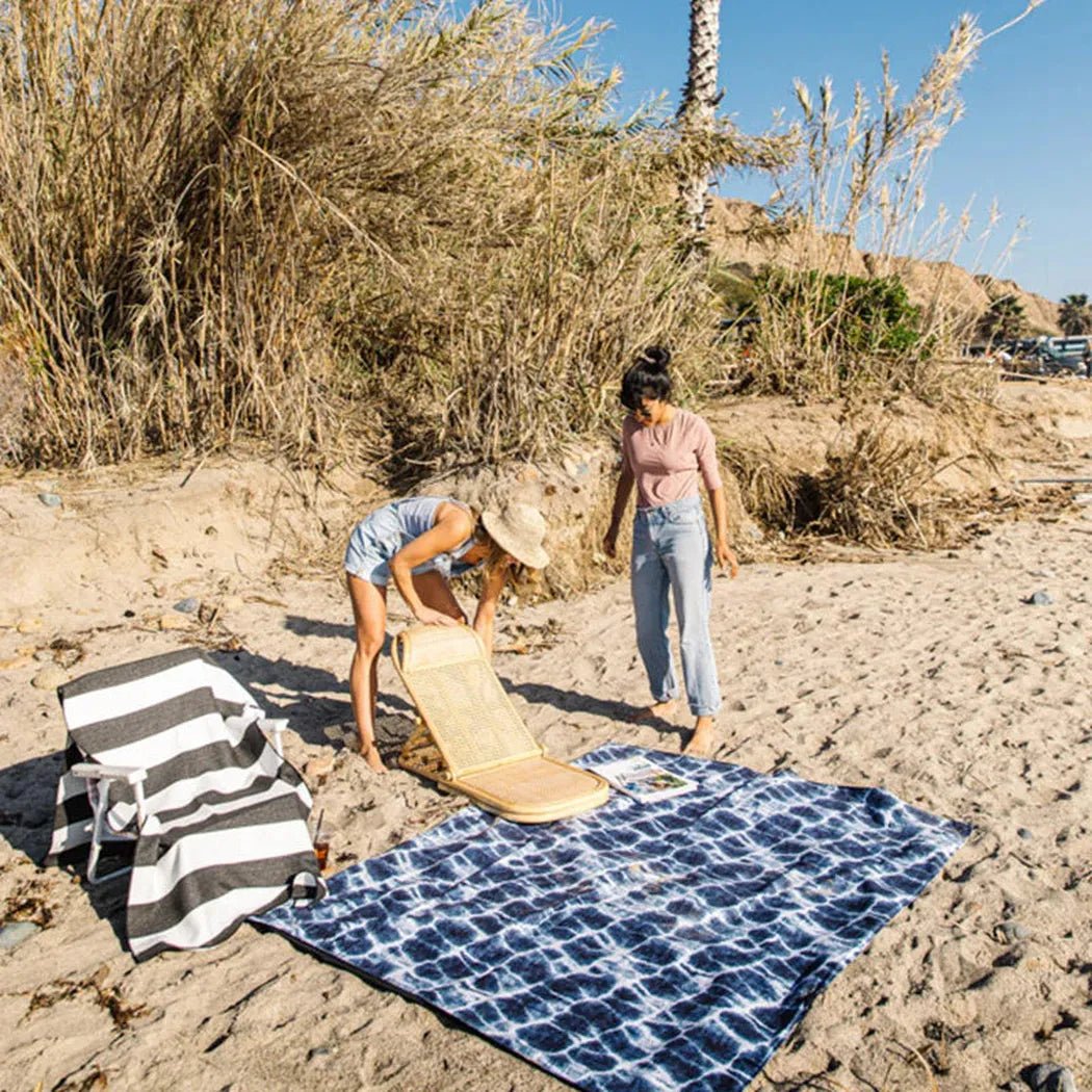 A woman sets up a Nomadix Festival Blanket in Baja Aqua on a sandy beach with tall grass and a palm tree in the background.