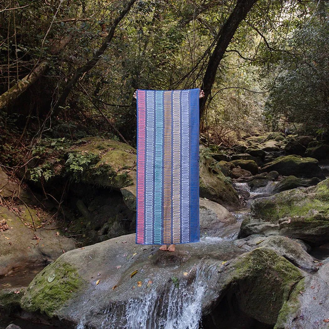A person stands on a mossy rock by a stream holding the Nomadix Original Towel in Agua Blue, with a forest background.