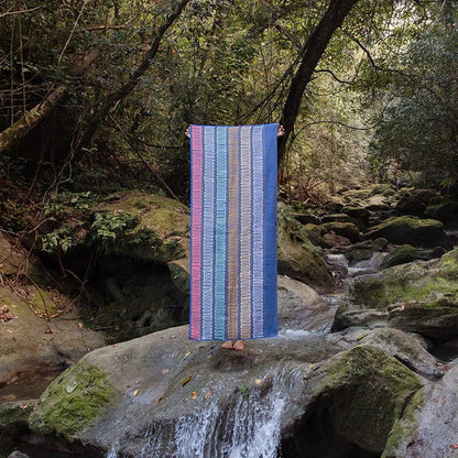 A person stands on a mossy rock by a stream holding the Nomadix Original Towel in Agua Blue, with a forest background.