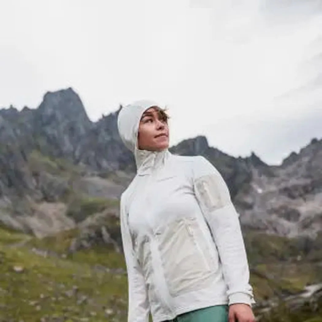 A woman wears the Norrona Women's Falketind Warm 2 Stretch Hood in Indigo Night while standing in front of rocky mountains.