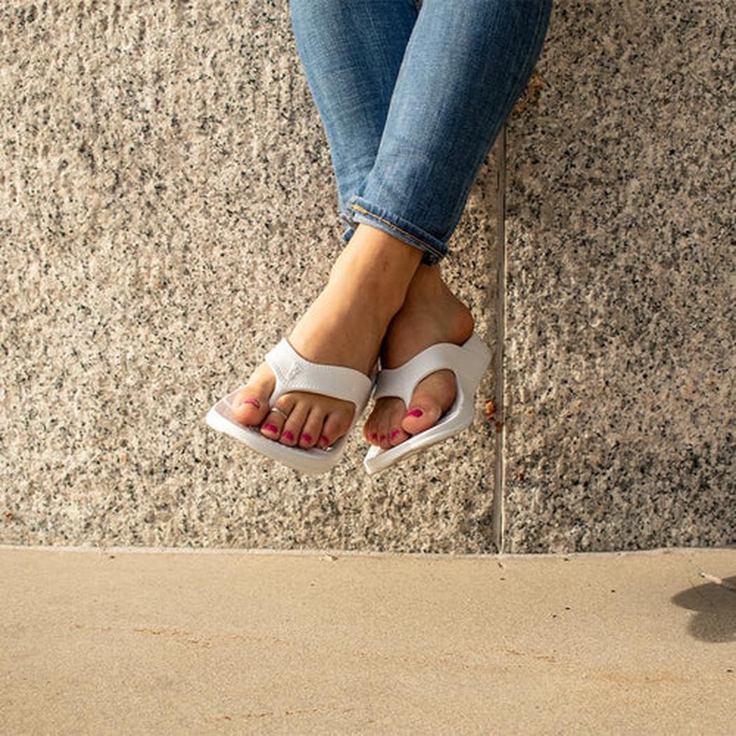 A woman wearing blue jeans and white NuuSol Womens Cascade Flip Flops with pink nail polish stands against a textured wall.